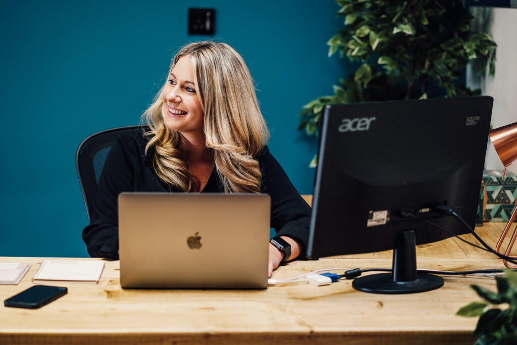 Anna, Behaviour Change expert at MacMartin, sat at her desk in front of a laptop in front of dark teal wall. Anna is looking to the side and smiling.