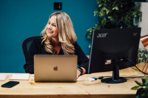 Anna, Behaviour Change expert at MacMartin, sat at her desk in front of a laptop in front of dark teal wall. Anna is looking to the side and smiling.